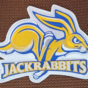 Puede incluir: Parche bordado con un jackrabbit amarillo con detalles azules y blancos. El jackrabbit está saltando, con la palabra "JACKRABBITS" en letras azules y amarillas debajo. El parche tiene un borde blanco.