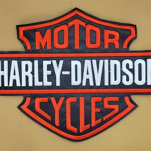 Op de afbeelding: Een zwart, oranje en wit Harley-Davidson Cycles-patch. De patch toont het Harley-Davidson-logo met de woorden "MOTOR", "HARLEY-DAVIDSON" en "CYCLES" in oranje en witte letters.