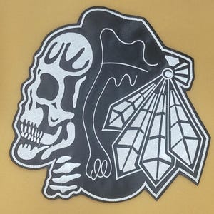 Könnte beinhalten: Schwarz-weißer Aufnäher mit einem Totenkopf und einem indianischen Kopfschmuck. Der Totenkopf befindet sich links, der Kopfschmuck rechts. Der Aufnäher hat einen genähten Rand.