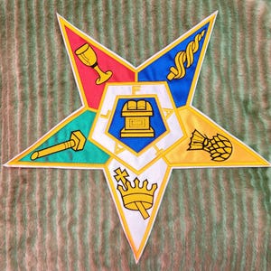 Pode incluir: Um emblema colorido em forma de estrela com uma borda branca. A estrela é dividida em segmentos de vermelho, azul, verde, amarelo e branco, cada um contendo um símbolo. O centro apresenta um design azul e branco com as letras "F A I T H".