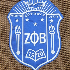 May include: A blue shield-shaped embroidered patch featuring white Greek letters ZΦB in the centre. Above the letters are four stars and a dove. Below are two torches on the sides and an open book with the year 1920. The text "πάντα ἐργασία νίκα" is arched across the top.