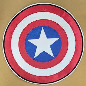 Puede incluir: Un escudo redondo con un diseño rojo, blanco y azul. El escudo presenta un anillo exterior rojo, un anillo interior blanco y un centro azul con una estrella blanca. Un borde negro perfila el escudo.