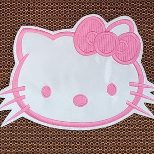 Peut inclure: Écusson brodé blanc et rose représentant le visage d'un chat de dessin animé, avec un nœud rose, des moustaches et des traits du visage. Le patch a un contour rose et est posé sur un fond marron.