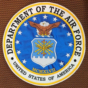 以下が含まれることがあります： アメリカ空軍のエンブレムが刺繍された円形パッチ。青色の中心部には金の縁取り、白い星、そして「DEPARTMENT OF THE AIR FORCE」と「UNITED STATES OF AMERICA」の文字が施されています。中央には鷲と盾が描かれています。