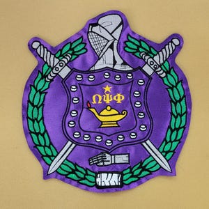 Könnte beinhalten: Ein lila Aufnäher mit einem Schilddesign, das eine goldene Lampe, griechische Buchstaben und einen Stern zeigt. Zwei silberne Schwerter kreuzen sich hinter dem Schild, umgeben von grünen Lorbeerzweigen. Der Aufnäher enthält die Buchstaben "AKA" unten.
