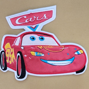 以下が含まれることがあります： 映画「カーズ」のライトニング・マックィーンが描かれた赤い刺繍ワッペン。ワッペンは白い縁取りがあり、車の上の看板に赤い文字で「Cars」と書かれています。車は青い目と笑顔の口をしています。