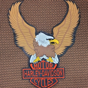 May include: Embroidered patch featuring a detailed eagle with outstretched wings, in shades of brown, tan, and black. The eagle is perched above a red and black Harley-Davidson Cycles logo, with the words "MOTOR HARLEY-DAVIDSON CYCLES".