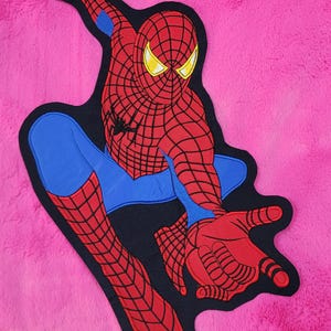 May include: A Spider-Man applique in red, blue, and black, set against a pink backdrop. The design depicts the superhero in a dynamic pose, with outstretched arms. The applique features the web pattern and yellow eyes.