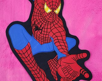 Könnte beinhalten: Ein rotes, blaues und schwarzes Spider-Man-Applikation auf pinkfarbenem Hintergrund. Die Applikation zeigt den Superhelden in einer dynamischen Pose mit ausgestreckten Armen. Das Design umfasst das Netzmuster und gelbe Augen.