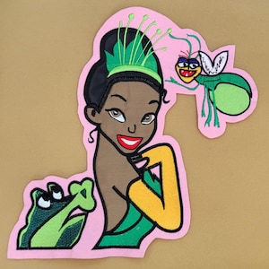 Może przedstawiać: Haftowana naszywka przedstawiająca postać z kreskówki, kobietę o ciemnej karnacji, ubraną w zieloną suknię i tiarę. Żaba i świetlik są również zawarte w projekcie, wszystko na różowym tle.