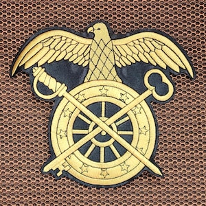 May include: Embroidered patch featuring a gold eagle with outstretched wings above a wheel design. A sword and key cross the wheel. The patch has a black background and gold stitching.