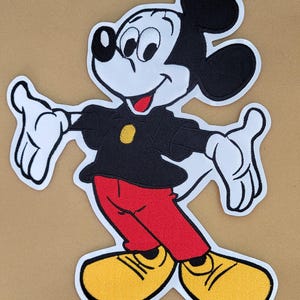 May include: Embroidered patch featuring Mickey Mouse in a classic pose. Mickey is depicted with black ears, a black shirt, red pants, and yellow shoes. The patch has a white border and is set against a tan background.