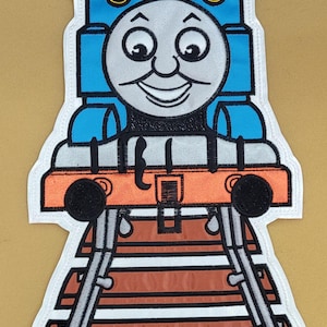 Op de afbeelding: Geborduurde patch met een cartoon trein met een lachend gezicht. De trein is voornamelijk blauw, met een grijs gezicht, oranje accenten en zwarte wielen. De patch bevat spoorrails.