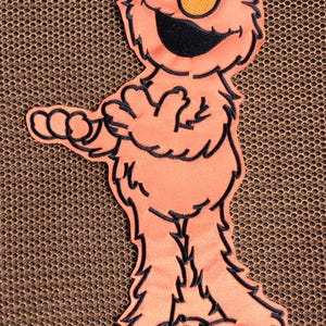May include: A coral-colored patch featuring the Sesame Street character Elmo. The patch has a black outline and depicts Elmo with a smiling face, white eyes, and an orange nose. The background is a brown mesh.