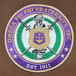 May include: A circular embroidered patch featuring the Omega Psi Phi Fraternity, Inc. emblem. The design includes a purple border with gold text, a central shield with a lamp and Greek letters, crossed swords, a helmet, and green laurel wreaths.