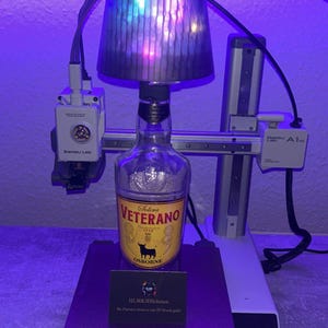 May include: A clear glass bottle of Solera Veterano brandy with a yellow label and a bull logo, placed on a 3D printer. The printer has a silver frame and a purple base. A lampshade is mounted above the bottle.
