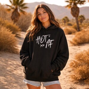 Op de afbeelding: Zwarte hoodie met de tekst "HOT AF" in het wit, met een capuchon met trekkoord en een voorzak. De hoodie wordt gedragen in een woestijnachtige omgeving met palmbomen en droge struiken op de achtergrond.