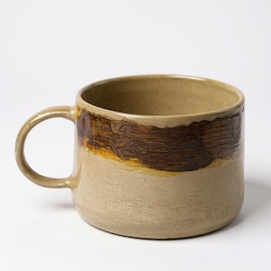 Peut inclure: Une tasse en céramique faite à la main avec une base arrondie et une grande anse. La tasse présente un design bicolore, avec une moitié inférieure beige clair et une bande texturée marron foncé sur la partie supérieure.