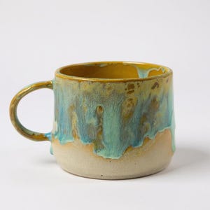 Puede incluir: Una taza de cerámica hecha a mano con un esmalte único. La taza presenta un interior y un asa amarillos, con una mezcla de colores azules, verdes y beige que caen por el exterior. La taza tiene una forma redondeada y un asa robusta.