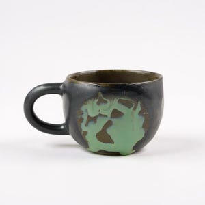 May include: A handmade ceramic mug with a black exterior and a green, abstract design. The interior rim is a dark brown. The mug has a rounded shape and a curved handle, perfect for enjoying coffee or tea.
