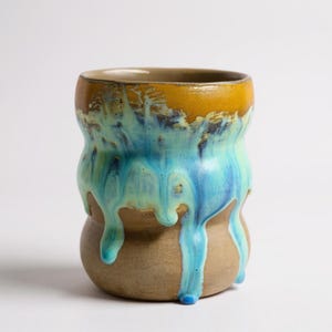 Könnte beinhalten: Eine handgefertigte Keramiktasse mit einem einzigartigen Design. Der obere Rand ist senfgelb und geht in eine tropfende türkisfarbene und blaue Glasur über einen beigefarbenen Sockel über. Die Tasse hat eine bauchige Form und eine glatte, glänzende Oberfläche.