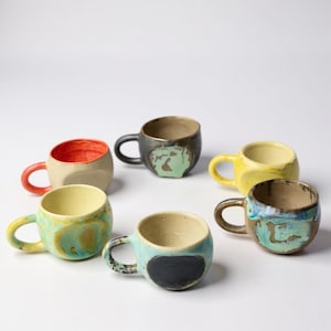 Puede incluir: Una colección de tazas de cerámica hechas a mano en varios colores y diseños. Las tazas presentan diseños únicos, incluyendo un interior rojo, un exterior negro con detalles verdes y un remolino turquesa y amarillo. Cada taza tiene un asa pequeña.