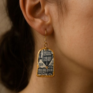 Könnte beinhalten: Ein goldfarbener Ohrring mit einem rechteckigen Anhänger mit einem schwarz-weißen architektonischen Design. Der Anhänger hat einen goldenen Rand und einen Haken für das Ohr. Der Ohrring wird am Ohr einer Person getragen.