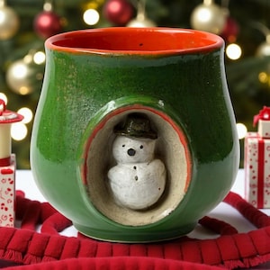 Peut inclure: Une tasse en céramique verte avec un motif de bonhomme de neige. L'intérieur est rouge et l'extérieur vert brillant. Un petit bonhomme de neige est niché dans une découpe circulaire à l'avant. Décoration de vacances festive.