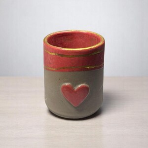 May include: A cylindrical ceramic container with a red heart and a red band with gold stripes. The container is gray below the red band and has a gold rim. The container is sitting on a light-colored wooden surface.