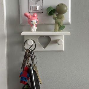 Puede incluir: Un llavero blanco montado en la pared con un recorte en forma de corazón y dos clavijas. Un juego de llaves cuelga debajo, con una figura de Lego de Spider-Man adjunta. Una figura rosa de My Melody y una figura de lámpara verde se sientan en un estante arriba.