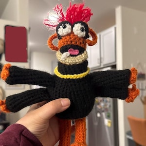 May include: A handmade crochet doll with a whimsical design. The doll features a black body, orange legs and arms, and a head with orange and tan details, large googly eyes, and a red mohawk. A yellow collar accents the neck.