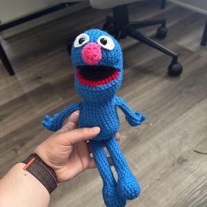 May include: A handmade blue crochet doll with a pink nose, red mouth, and white-rimmed eyes. The doll has arms and legs and is held in a person's hand. The doll is a character from Sesame Street.