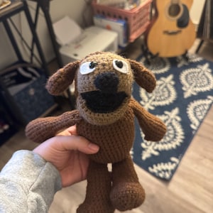 May include: A handmade brown crochet dog doll with white eyes and a black nose and mouth. The dog has floppy ears, arms, and legs. The doll is held in a person's hand.