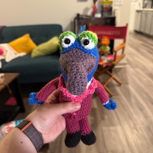 Puede incluir: Una marioneta de ganchillo hecha a mano al estilo de un personaje de los Muppets. La marioneta tiene una cabeza azul con ojos verdes y blancos, un pico morado y un cuello rosa. El cuerpo es de color magenta oscuro con pies negros y manos azules.