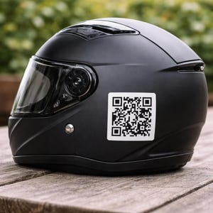 May include: A matte black motorcycle helmet with a clear visor and a QR code sticker. The helmet is sitting on a wooden surface, with a blurred green background.