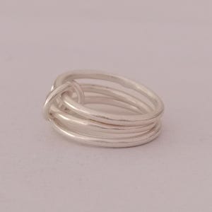 Könnte beinhalten: Ein silberner Ring mit einem Knotendesign. Der Ring hat mehrere Bänder, die übereinander gestapelt sind und ein einzigartiges, strukturiertes Aussehen erzeugen. Der Ring besteht aus einem glänzenden, metallischen Material.