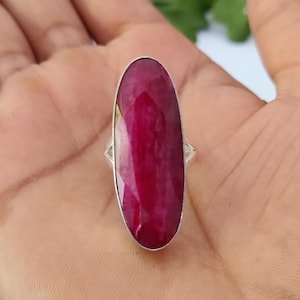 May include: A ring featuring a large, elongated oval-shaped, faceted deep magenta gemstone set in a simple silver-toned metal band. The stone has a translucent quality with visible internal inclusions and a smooth, polished surface texture.