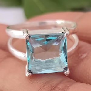 May include: A silver ring featuring a large, square-cut, light blue gemstone held by four prongs. The band is split into two thin, polished silver segments that meet at the base of the setting, creating a sleek and open design for the finger.