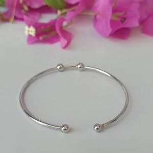 May include: A silver-colored, open-ended bangle bracelet with small, round ball accents. The bracelet is set against a backdrop of bright pink flowers and a white surface. The bracelet's design is simple and elegant.