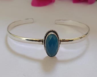 Könnte beinhalten: Ein silbernes Armreif-Armband mit einem ovalen, facettierten, teal-farbenen Edelstein in der Mitte. Der Edelstein ist in einem dekorativen, seilartigen Rahmen gefasst. Das offene Armband liegt auf einer weißen Oberfläche.