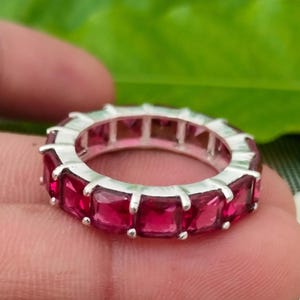 Peut inclure: Une bague alliance en argent ornée d'une rangée continue de pierres précieuses rouge foncé taillées en carré et serties par des griffes. La bague présente un design uniforme où chaque pierre est maintenue par quatre griffes métalliques.