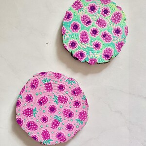 May include: Two round wooden coasters with hand-painted designs. One coaster has a pink background with a pattern of purple raspberries and green leaves. The other coaster has a teal background with a similar raspberry design.
