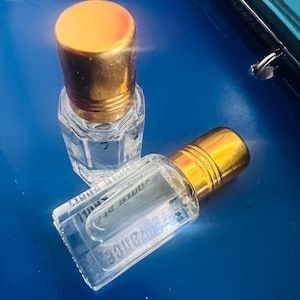 May include: Two small, clear glass perfume bottles with gold-colored caps. The bottles are filled with a clear liquid and are resting on a blue surface. One bottle is upright, while the other is lying on its side.