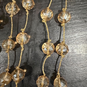 May include: A decorative garland featuring faceted, translucent amber-colored glass beads strung along a thin, metallic gold-colored cord. The beads are spaced out at regular intervals, each secured in place by small knots tied in the gold cord.