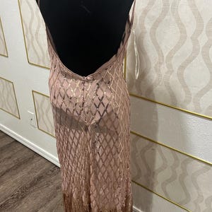 May include: A blush-coloured, floor-length dress with a low back and spaghetti straps. The dress features a diamond pattern of gold sequins and a fringe hem. The dress is displayed on a black mannequin.