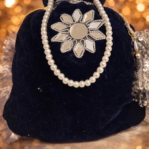 Puede incluir: Una bolsa de terciopelo azul marino con un collar de perlas sobre ella. La bolsa está decorada con un diseño de flor espejada. Una borla plateada cuelga de un lado. El fondo es de color dorado borroso.