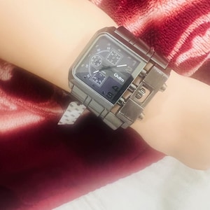 Könnte beinhalten: Eine große, quadratische Armbanduhr mit Gunmetal-Finish. Die Uhr verfügt über mehrere Hilfszifferblätter und den Markennamen "Oulm" auf dem Zifferblatt. Die Uhr wird mit einem passenden Metallarmband befestigt.