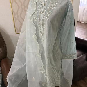 May include: A light blue embroidered tunic and matching sheer scarf. The tunic features intricate white floral embroidery around the neckline and sleeves. The scarf has a scalloped edge with matching embroidery. The outfit is displayed on a black mannequin.