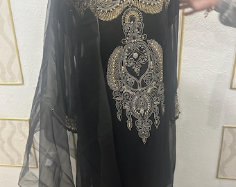 May include: Black dress with gold embroidery. The dress has detailed gold embellishments around the neckline, sleeves, and a central design. A sheer black shawl drapes over the shoulders. The dress is displayed on a mannequin.