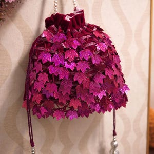 Könnte beinhalten: Burgunderfarbener Beutel mit Kordelzug, verziert mit glitzernden, blattförmigen Pailletten in Rosa- und Magentatönen. Die Tasche hat einen perlenbesetzten Griff und Quastenakzente, was sie zu einem eleganten Accessoire macht. Maße in Zentimetern.